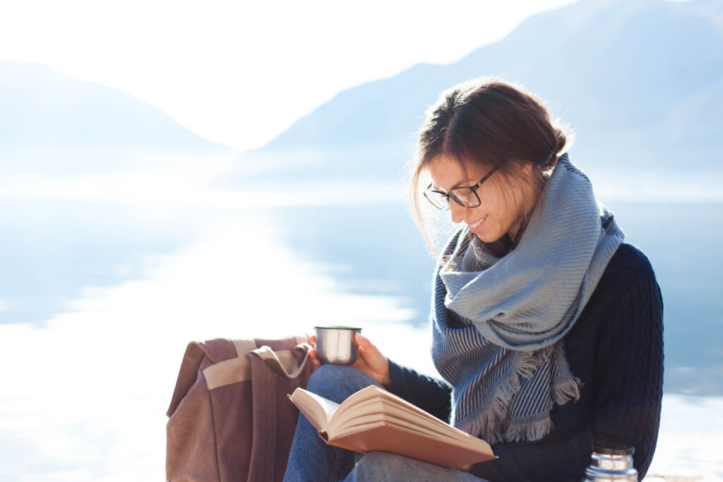 Young woman reading book at sea beach. Cozy winter picnic by morning mountains. Happy student